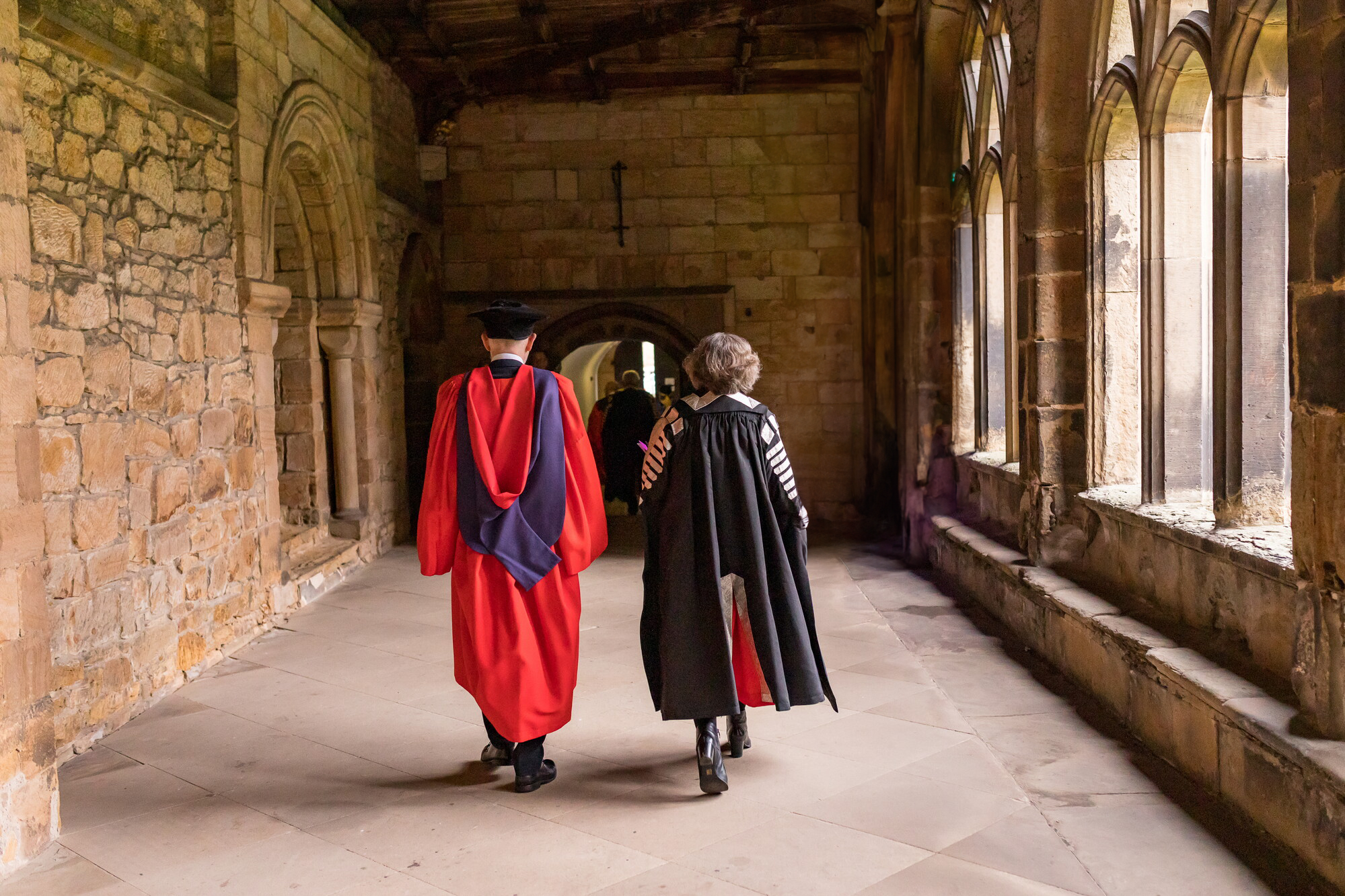 Two people in academic gowns walk along the cloisters