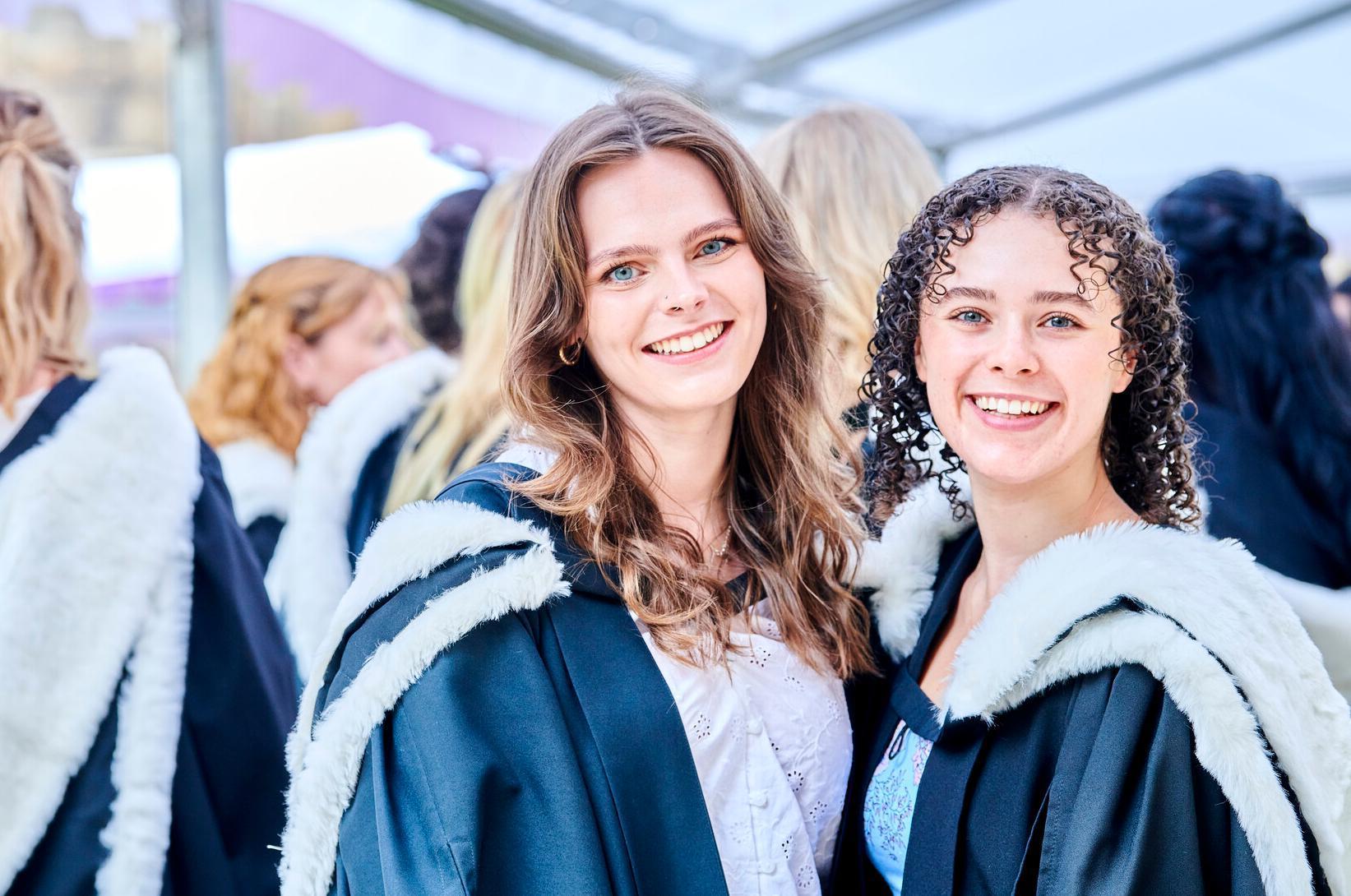 Two students in graduations gowns smiling in a crowd of gowned people