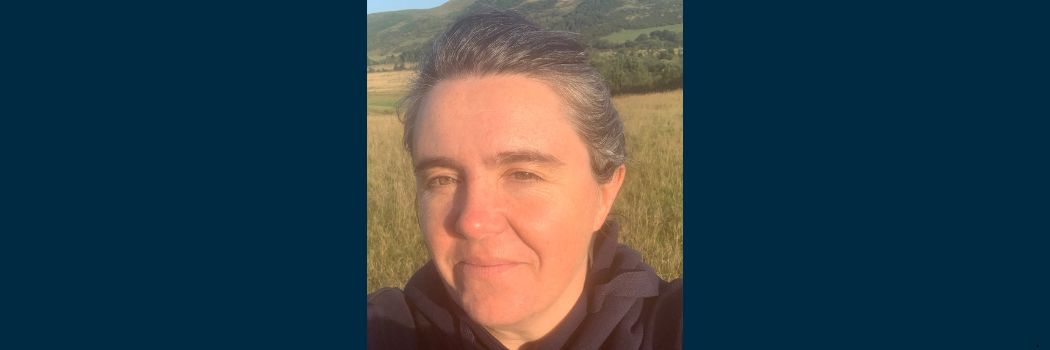 A head and shoulders close up photograph of Professor Sarah Semple. Sarah is smiling at the camera while standing in a field surrounded by countryside.