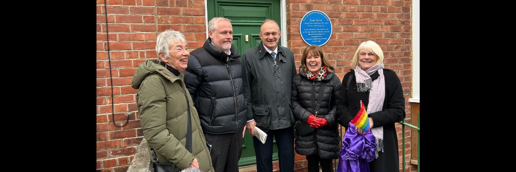 People standing in front of a house with a green door and a blue plaque, smiling
