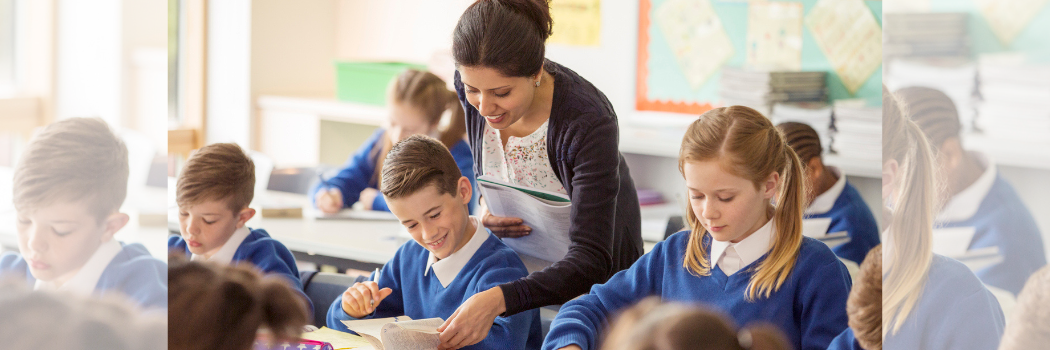 A female teacher in a classroom leaning over a schoolboy to turn a page of a book as they both smile