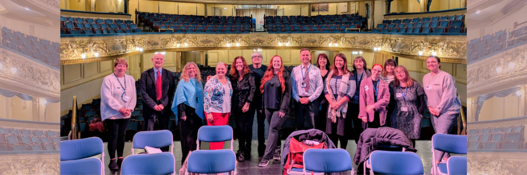 A group of adults dressed smartly standing on a theatre stage with the stalls behind them