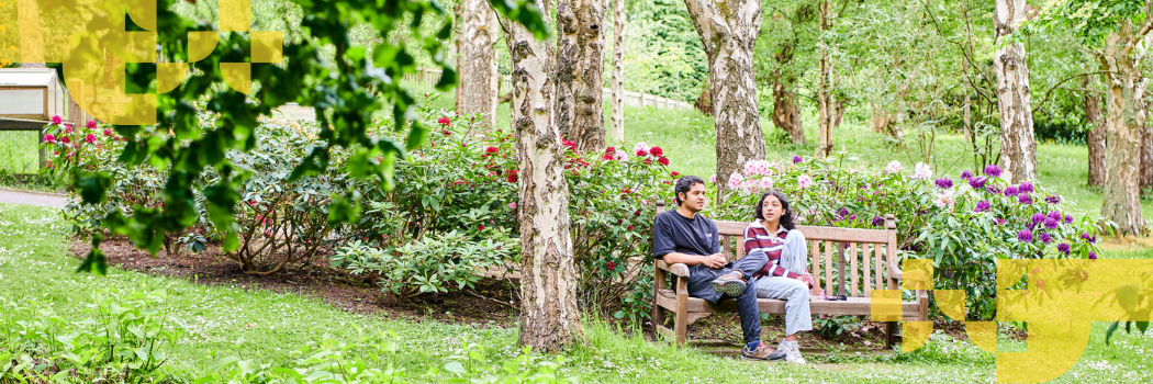 A male and female student sitting on a wooden bench surrounded by attractive parkland