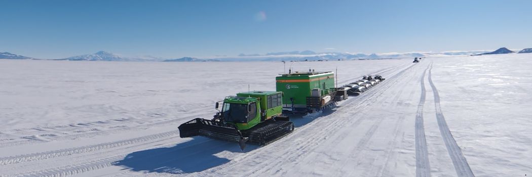 A snowplough drives across the Antarctic ice against a clear blue sky