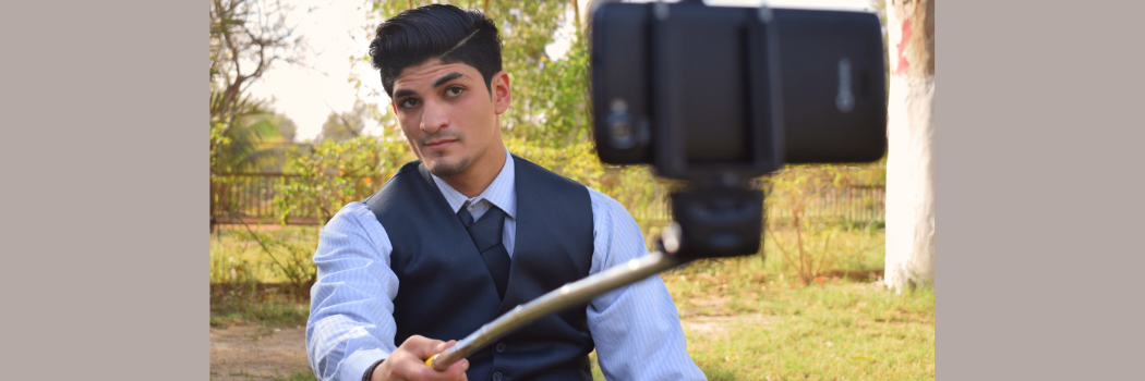 A man wearing a smart blue shirt and waistcoat poses for a selfie using a selfie stick