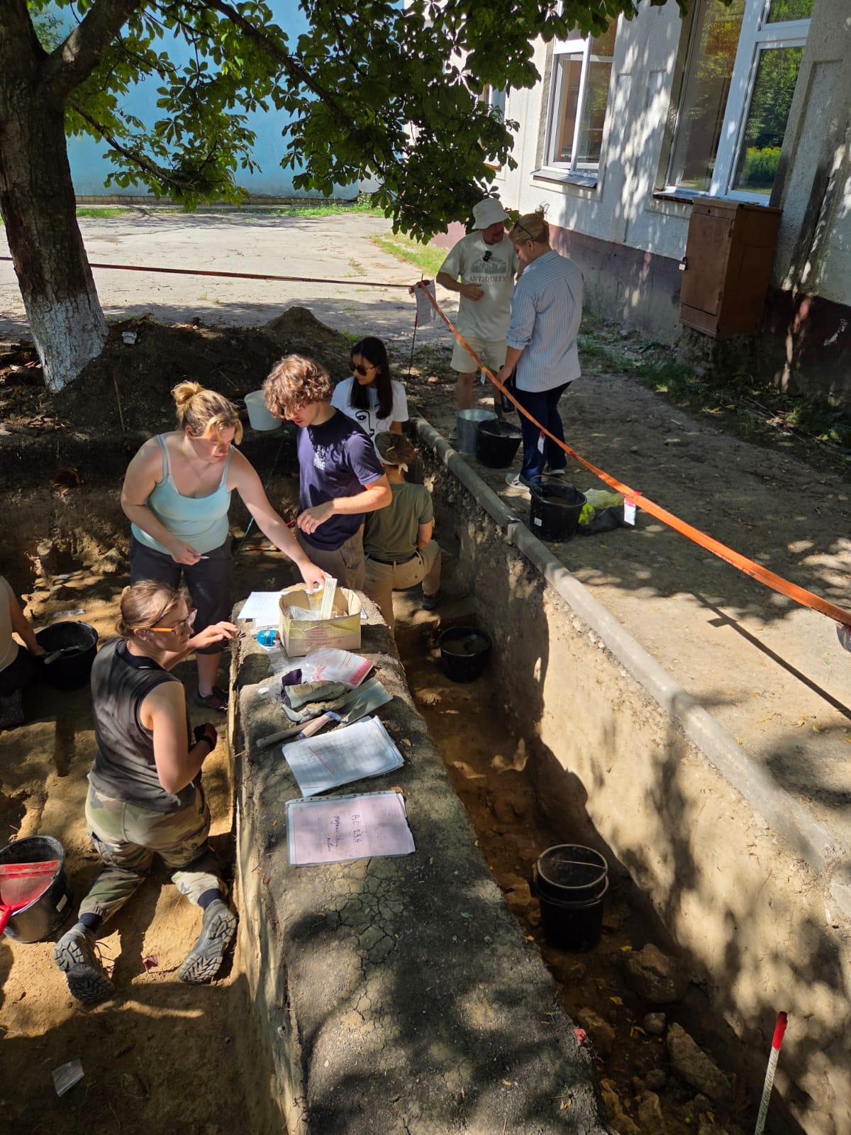 Five students working in an archaeological trench. On the side of the trench, Prof Paul Pettitt is talking to Deputy Ambassador Samantha Smith.