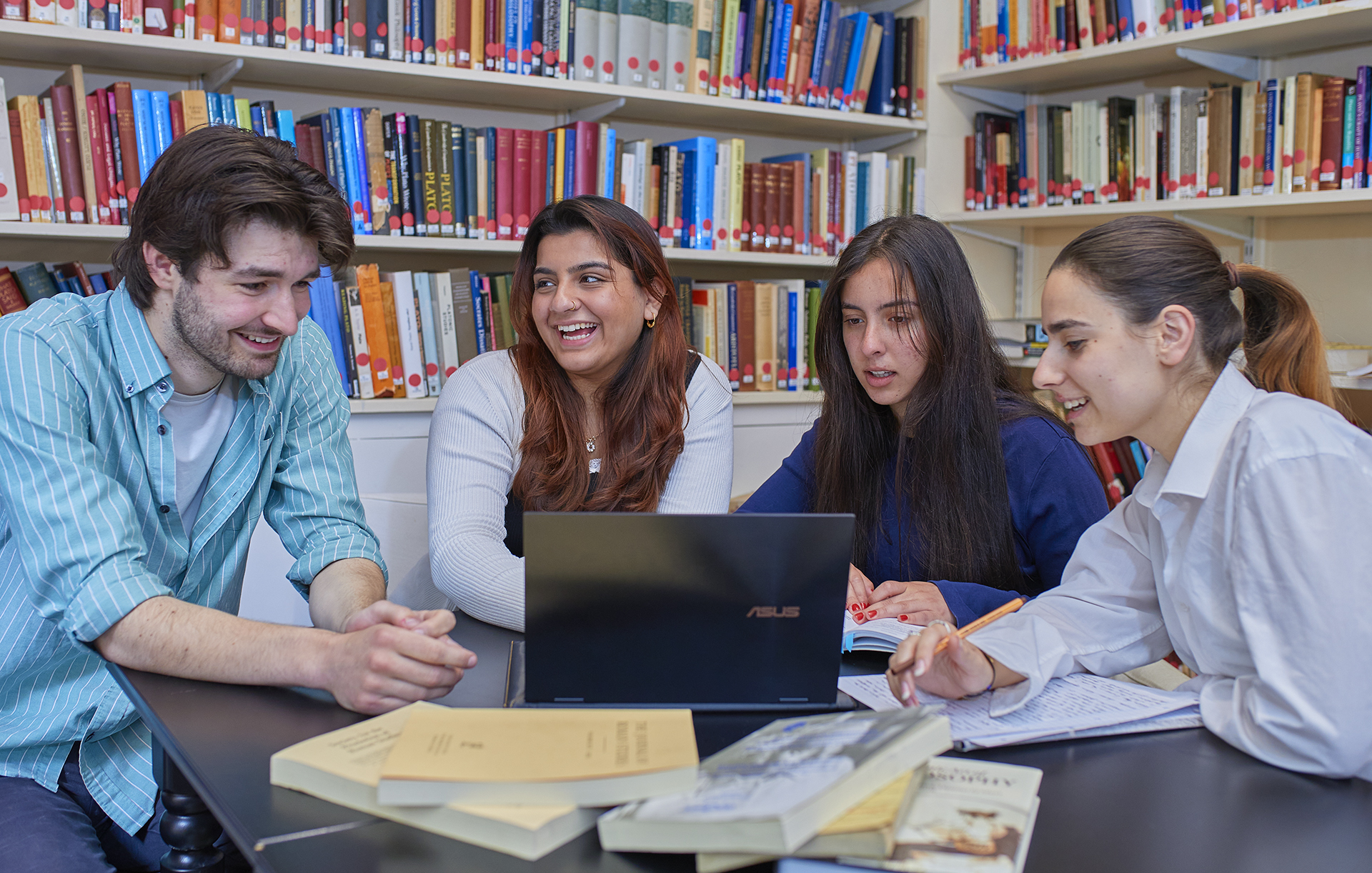 Four students sit at a table in a library with an open laptop, looking at each other and laughing