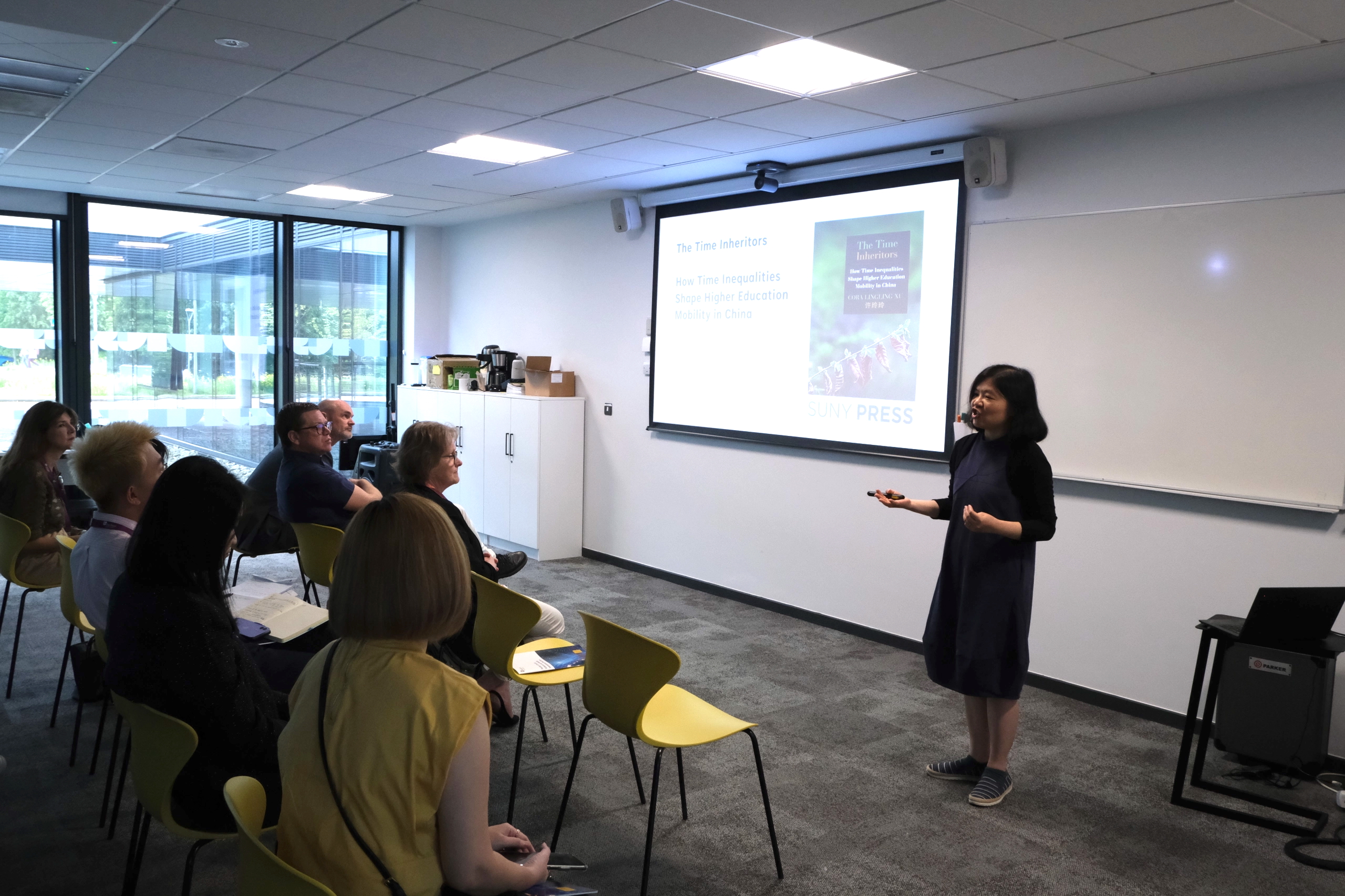 Dr Cora Lingling Xu stands at the front of a room, a PowerPoint presentation is projected on the wall behind her. Seated attendees are listening to her talk.