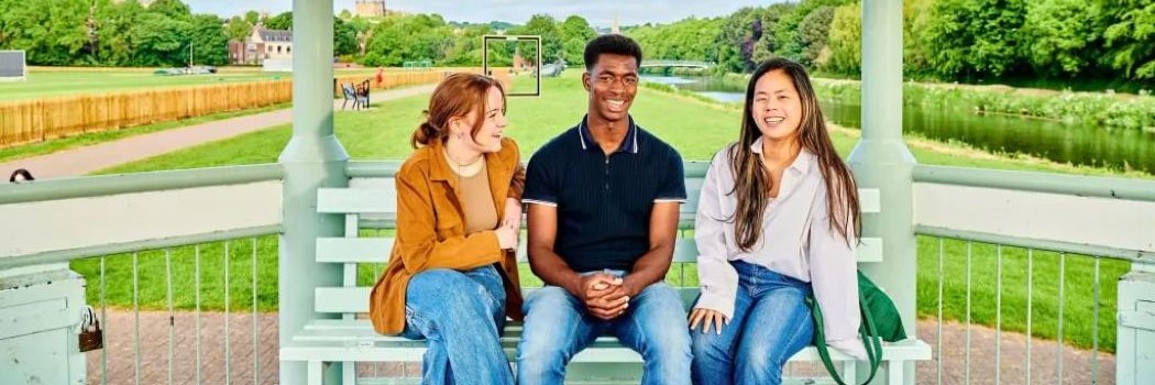Three students sat on a bench with a field behind them