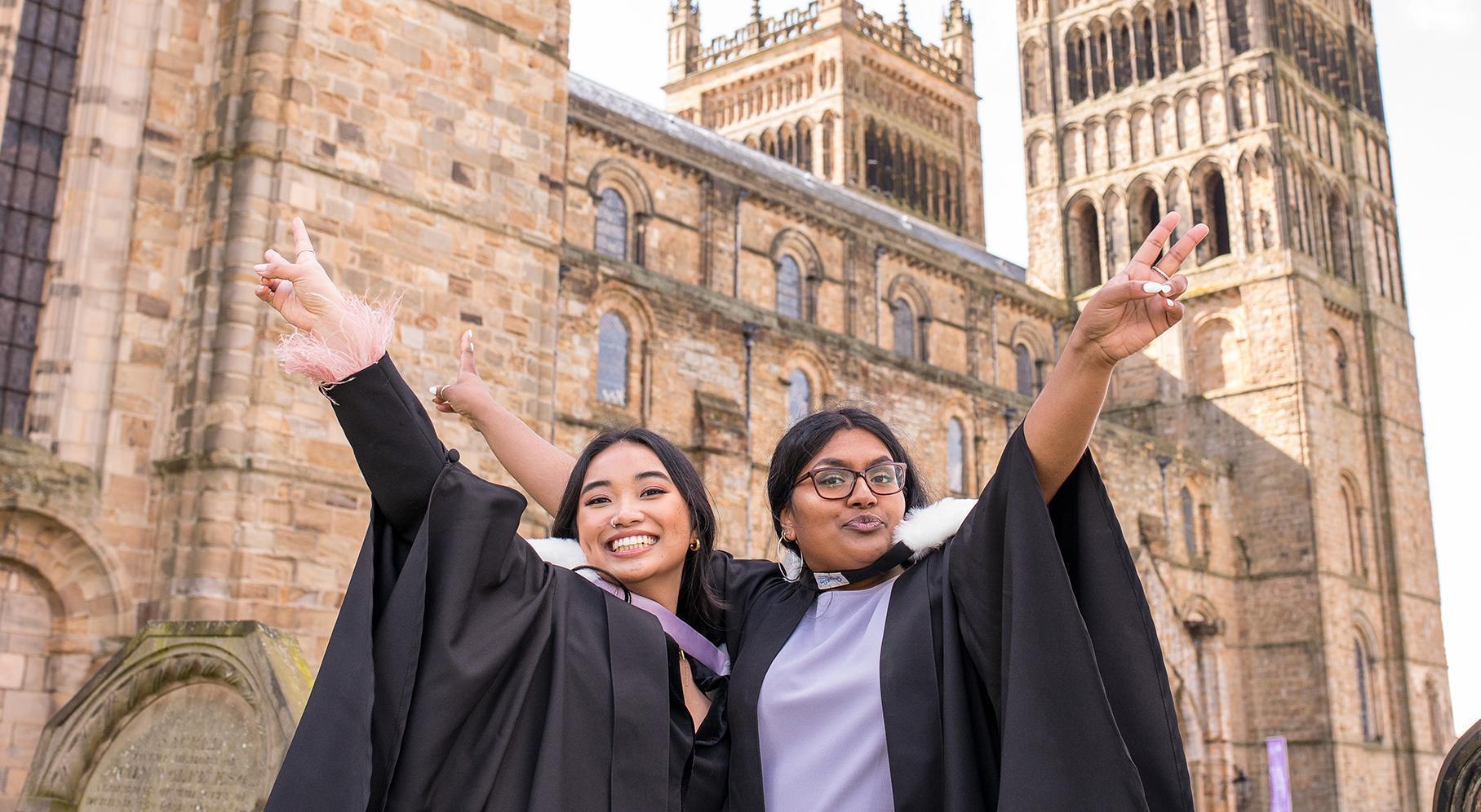 Two graduates in robes posing outside Durham Cathedral