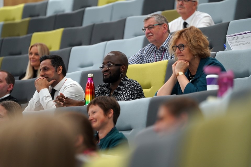 Men and women sit inside a modern looking lecture theatre. The camera is focused on three people who sit in the same row - they are all smiling and looking off camera