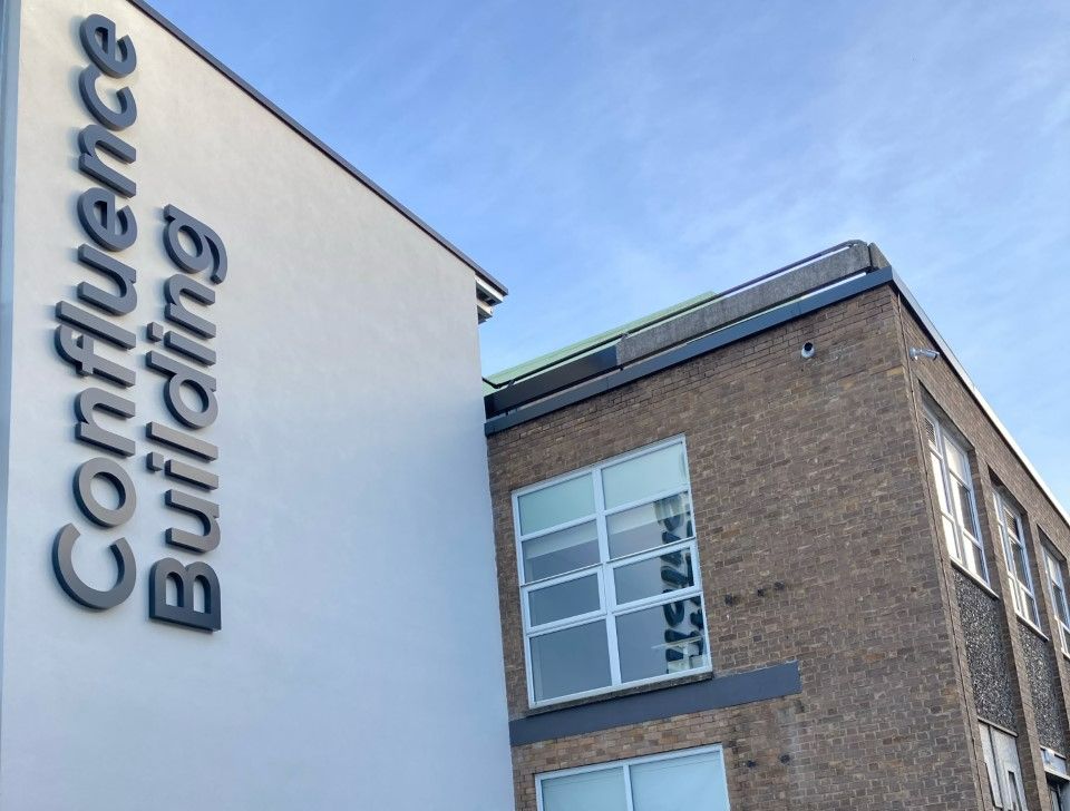 Vertical sign reading 'Confluence Building' next to a brown-bricked square building beneath blue skies.