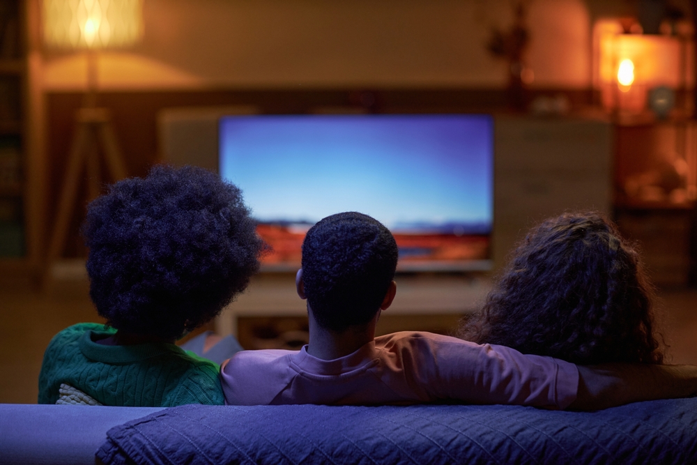 Photograph of three people sitting on a sofa watching the television, with atmospheric lighting and backs facing the camera.