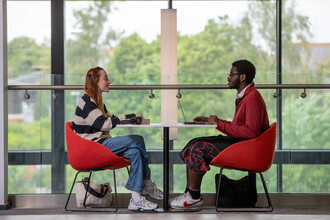 Two students sit opposite each other at a table. They are inside a modern building with lots of large windows