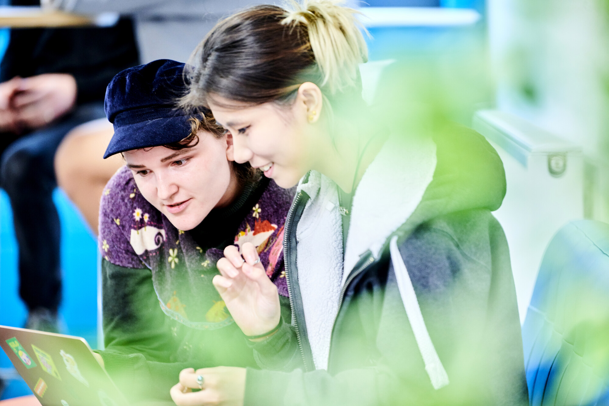 Two female students sit in a reception area, they are smiling and looking at their laptops