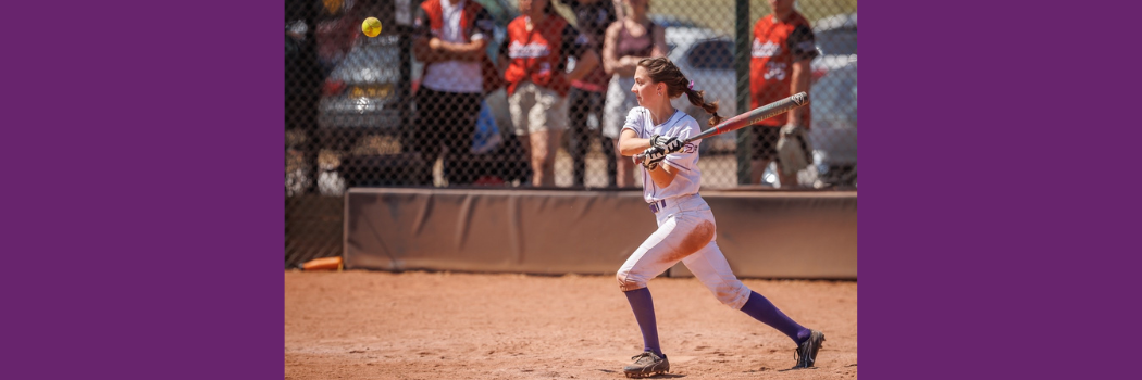 A young woman in baseball clothing and with her hair in a plait holds a baseball bat having just swung at a ball.