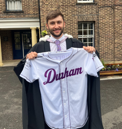 A young man wearing black graduation robes holds a white baseball shirt with Durham written across the front in purple.